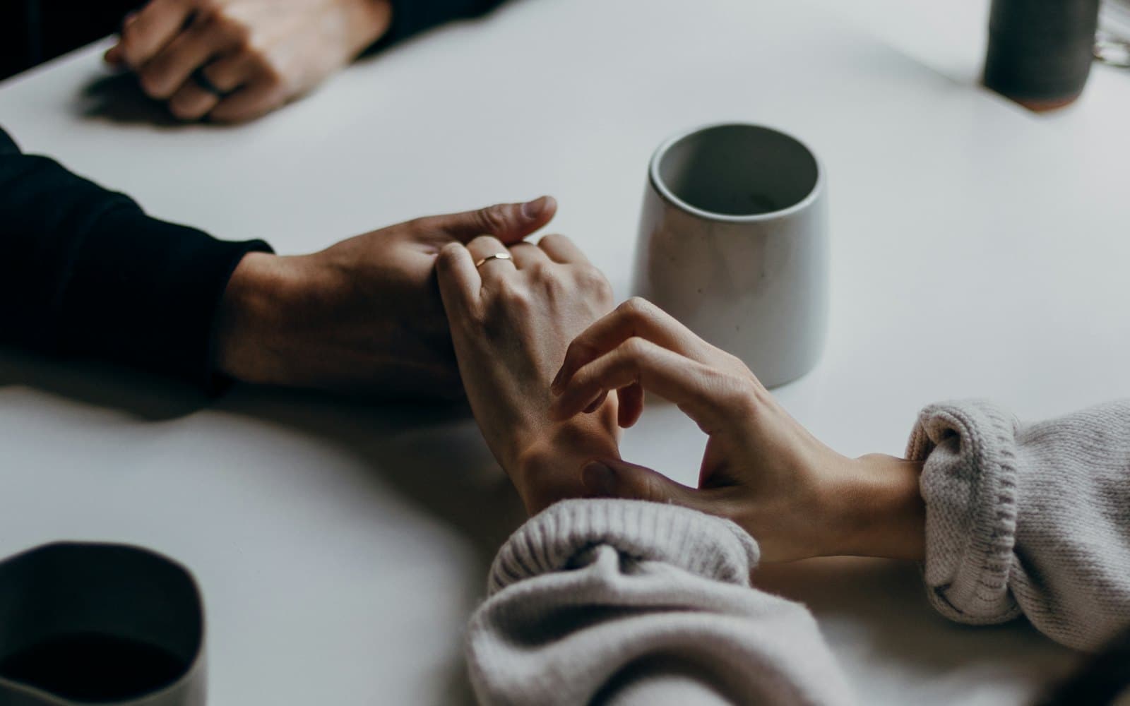 Two people sitting at a table with mugs, one hand resting on the other